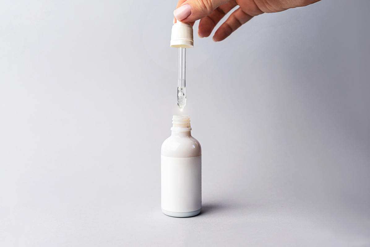 Hand of woman with pipette with drop of serum or hyaluronic acid or collagen moisturizer and white bottle on gray background.